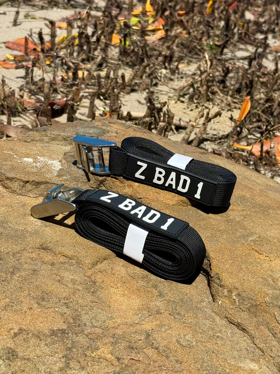 Two black ZBAD1 marine tension straps with stainless steel cam buckles coiled on a rock